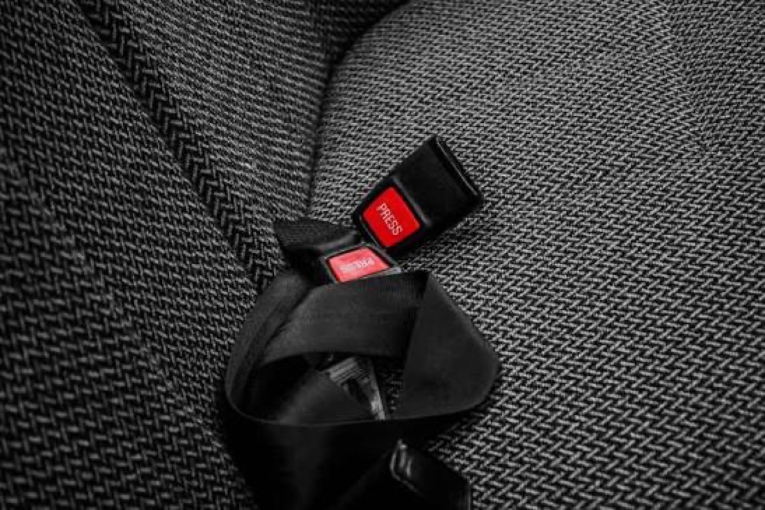 Detail of a black seatbelt buckle receptacle with a red release button set into a woven grey vehicle seat.