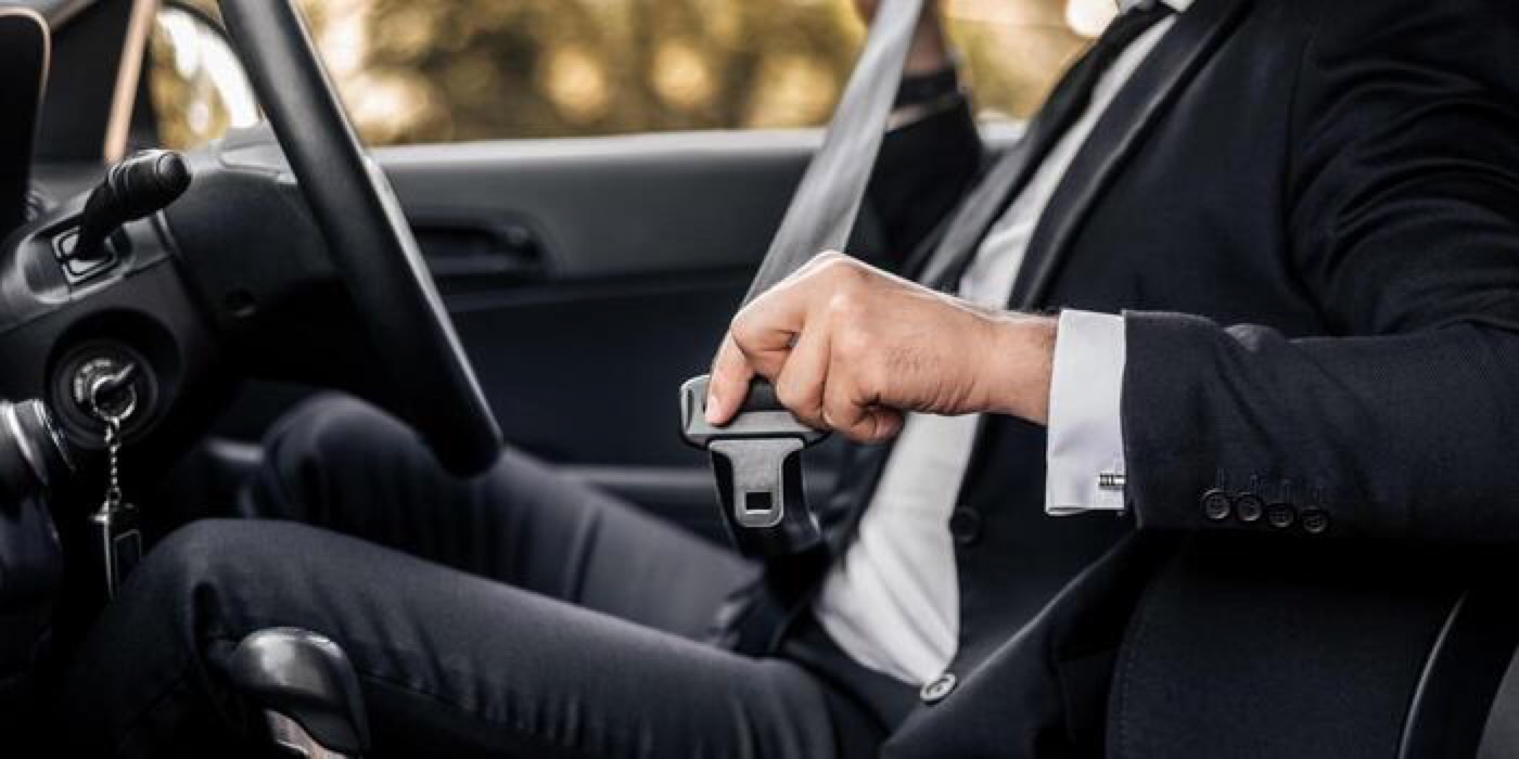 A person in a dark suit seated in a car, drawing the seatbelt across to fasten it before setting off.