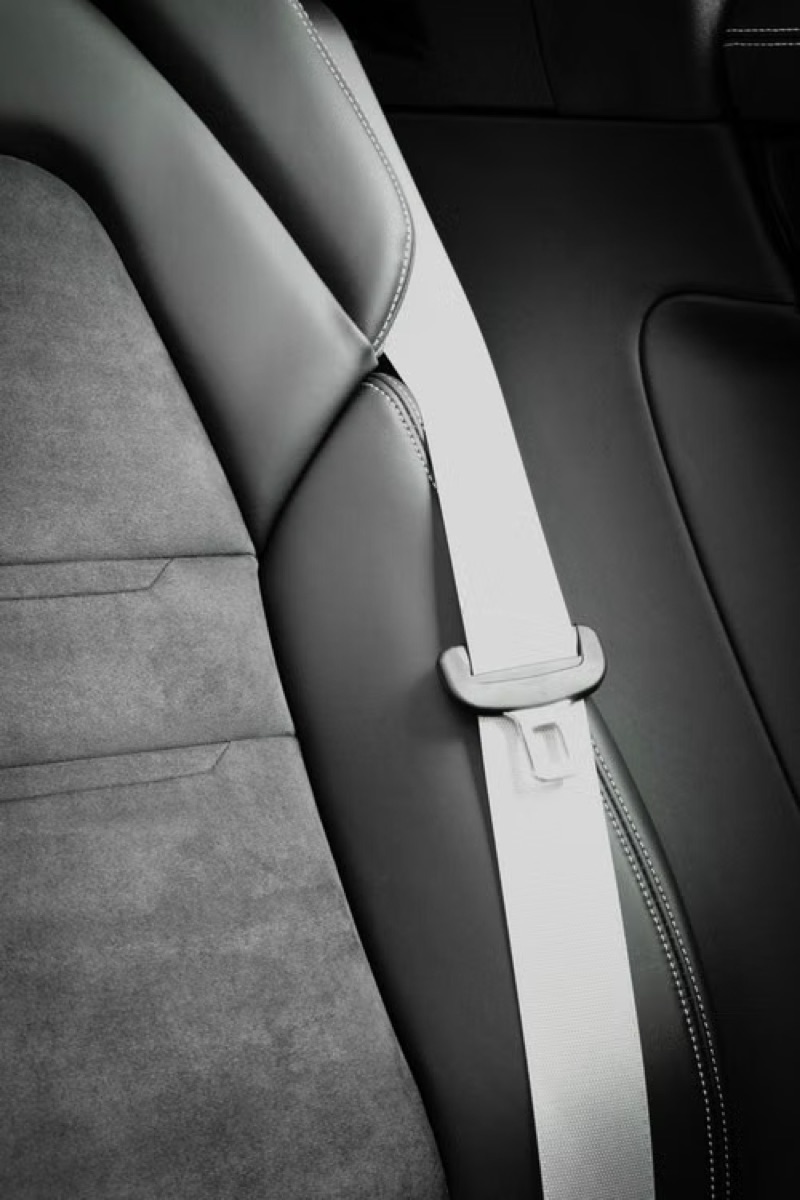 Light-coloured seatbelt webbing running across a dark vehicle seat, the latch plate clearly visible against the contrasting upholstery.